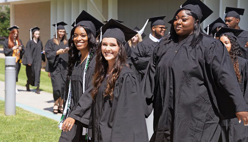 Two Meridian Community College graduates in caps and gowns smile while walking in a commencement procession.