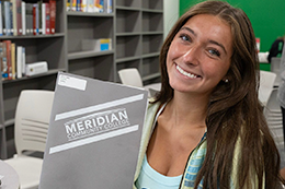 A female student smiles while holding up a gray MCC Orientation folder. 
