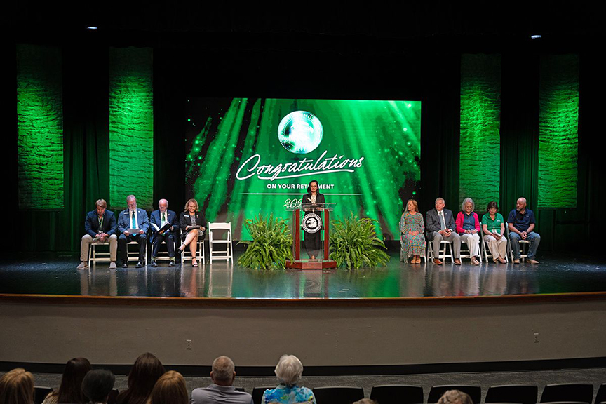 Dr. Leia Hill stands at a podium on stage during Meridian Community College’s Retiree Celebration ceremony, with honorees seated on both sides.